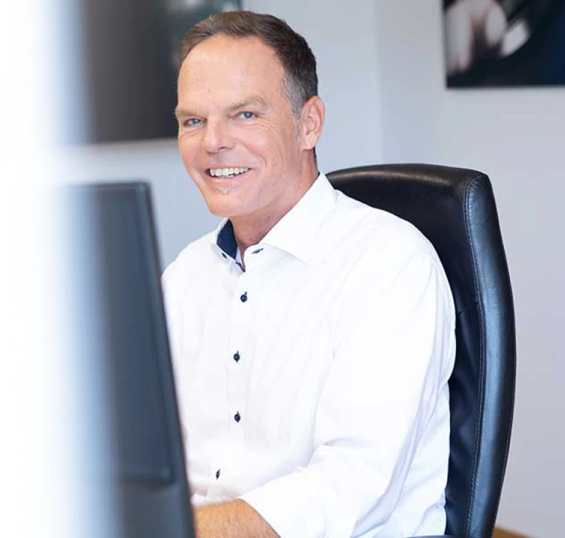 Lächelnder Steuerberater Stefan Hinterleitner im weißen Hemd am Büro-Arbeitsplatz zur Immobilienberatung.